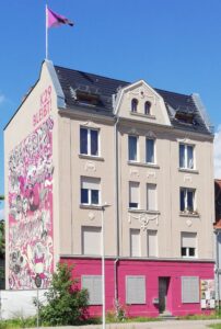 Frontansicht des Hausprojekt Zelle 79 in Cottbus. Auf der linken Seite ein großflächiges Graffiti. Auf dem Dach eine rosafarbene Fahne.