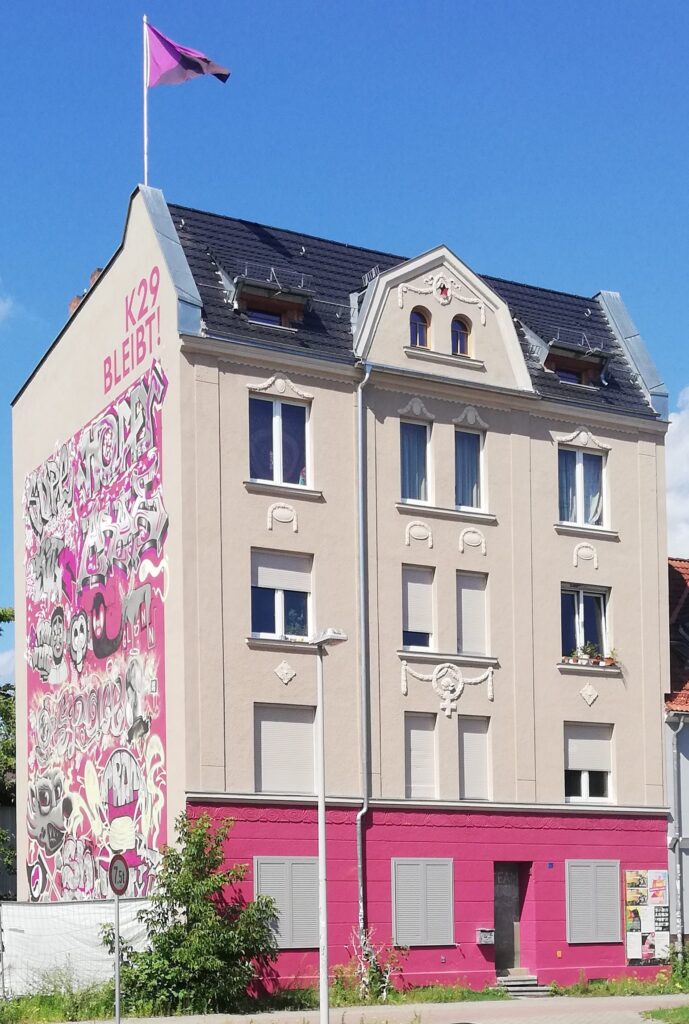Frontansicht des Hausprojekt Zelle 79 in Cottbus. Auf der linken Seite ein großflächiges Graffiti. Auf dem Dach eine rosafarbene Fahne.