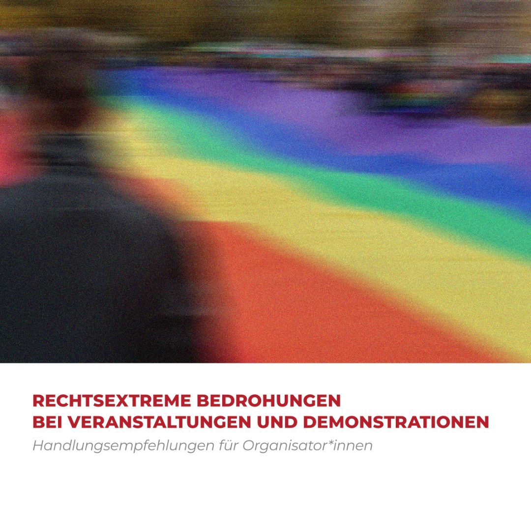 Verschwommen im Hintergrund eine große Regenbogenfahne, an den Seiten Menschen auf einer Versammlung.